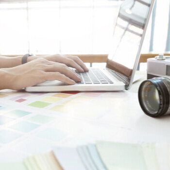 Hands typing on a laptop on a table covered in paint samples. Bright and sunny background