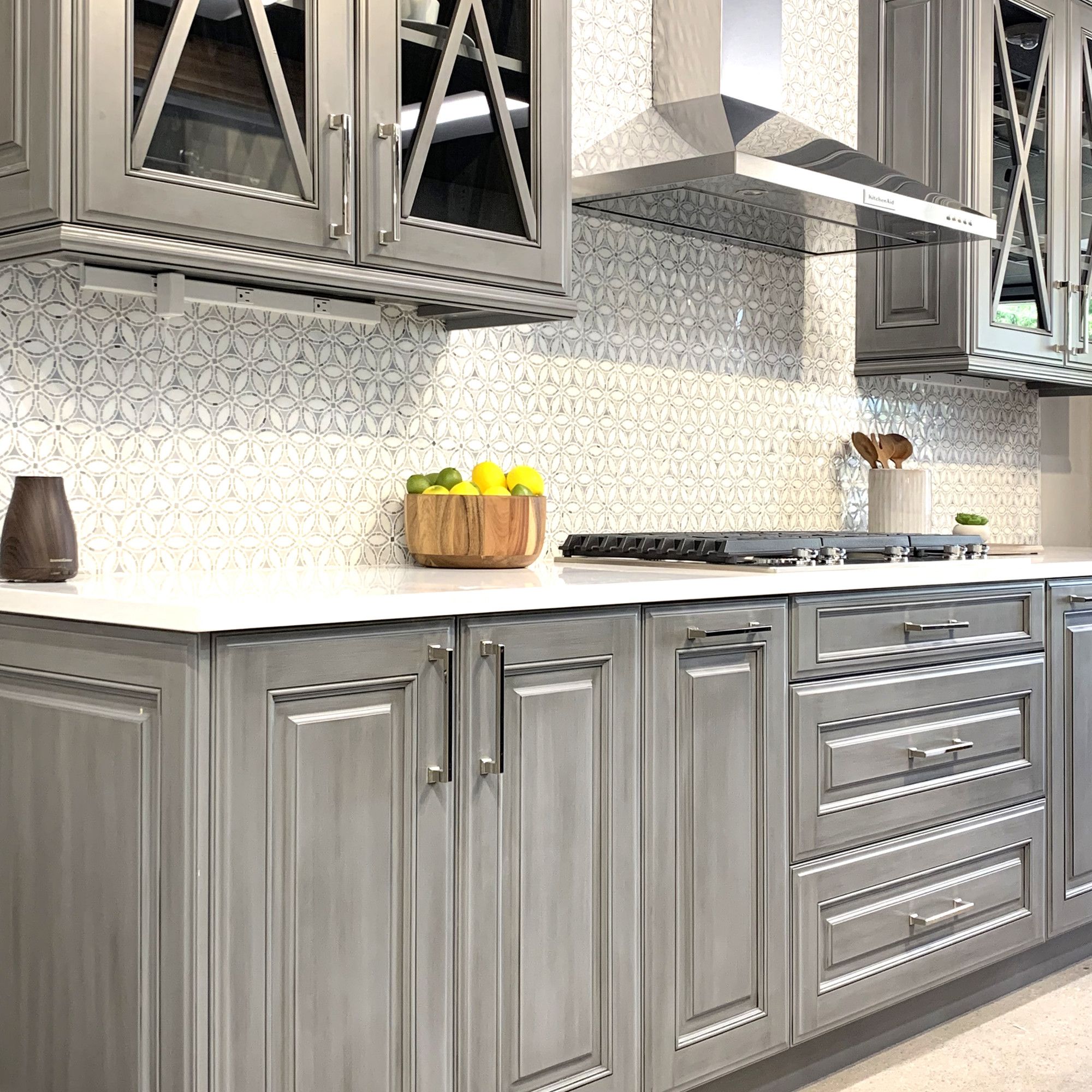 Kitchen with grey cabinets with silver handle, white countertops and a patterned backsplash. Upper cabinets have glass fronts with wood x design over glass
