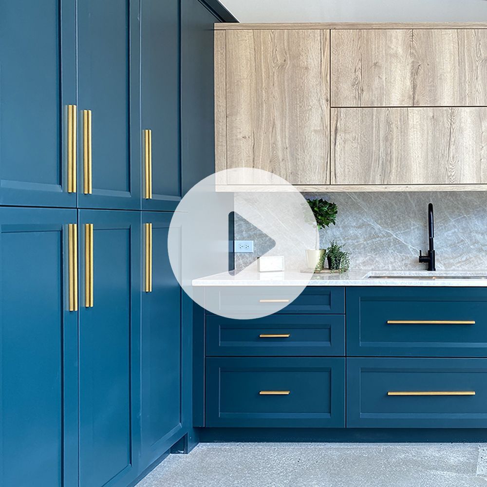 Kitchen with blue drawers and pantry doors that have gold handles. White marble countertops with a light wood upper cabinet and a granite backsplash. 