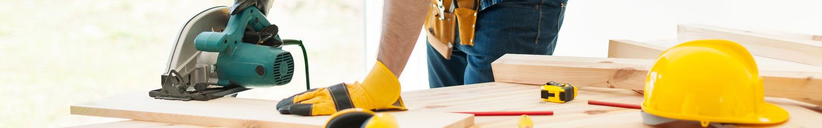 Construction worker using a circular saw to cut a board. They are wearing a tool belt, and work gloves. There is a safety hat and tools on the table next to where the board is being cut. 