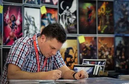 Artist at Kingcon behind his booth drawing while multiple prints of his art are displayed behind him
