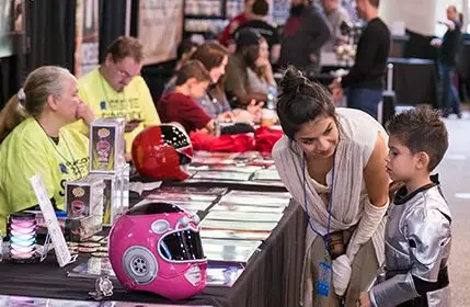 Vendor Hall at Kingcon with multipe comic books and art being sold at different booths that are in a line. A woman in a jedi cosplay is talking with a little boy, asking him what he would like to buy.