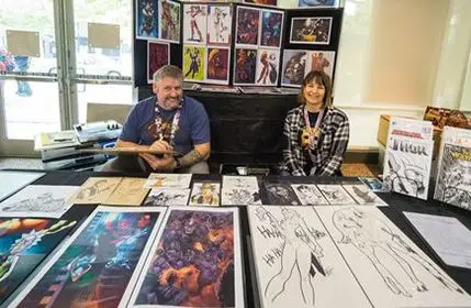 Two people a man and a woman sitting behind an artist's booth with their illustrations displayed on the table in front of them. 