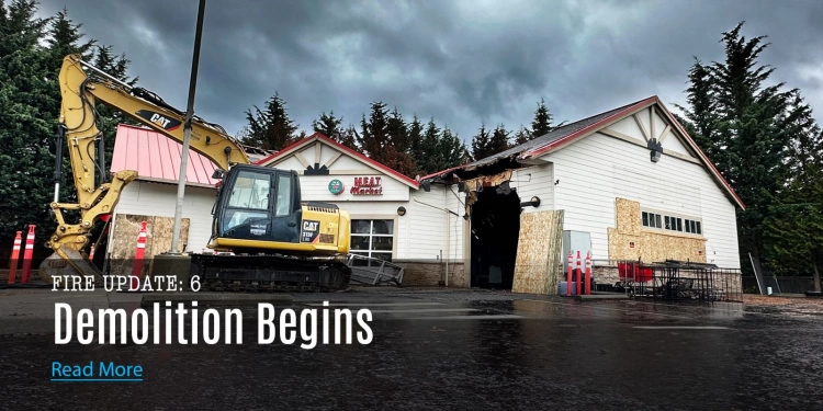 Construction equipment in front of the fire damaged Top of the Hill Quality Produce building with boarded-up windows and a 'Meat Market' sign, under a cloudy sky.