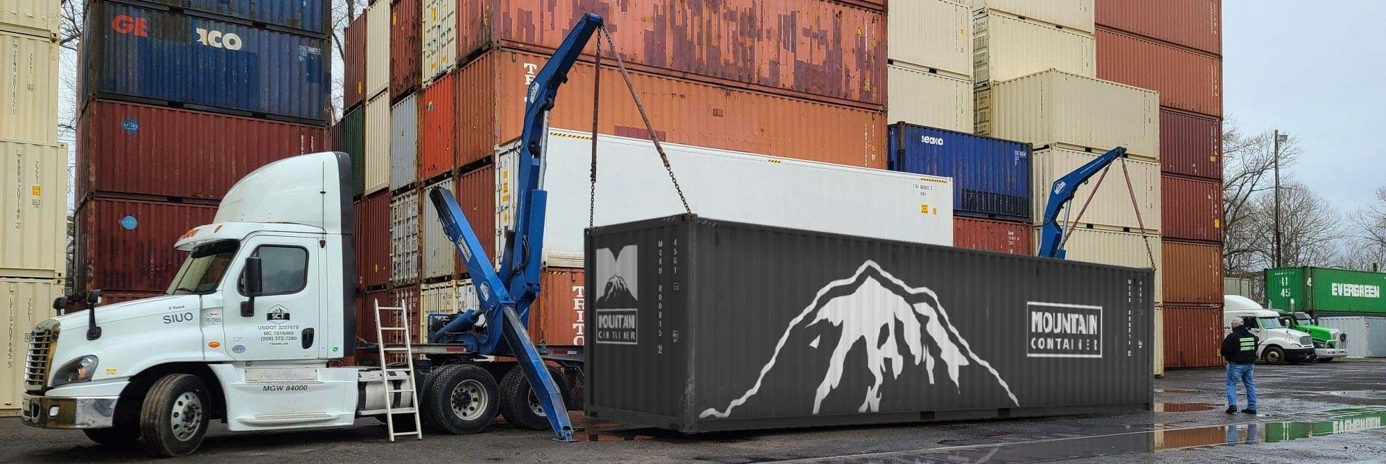 Grey Mountain Container Shipping container being loaded onto the back of a semi-truck using a side lifter in a shipping container lot. 