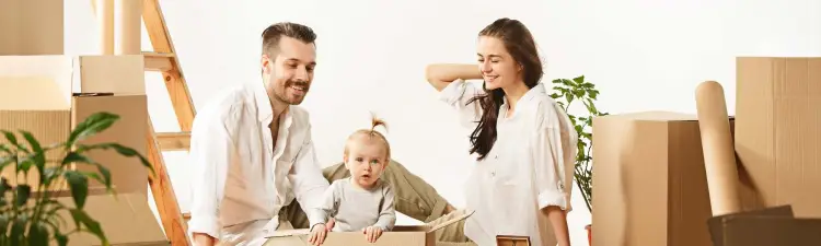 Smiling Man and Woman, looking at a toddler playing in a moving box in a room full of moving boxes.