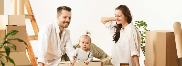 Smiling Man and Woman, looking at a toddler playing in a moving box in a room full of moving boxes.