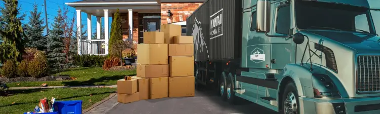 Branded shipping container on the back of a semi-truck in the driveway of a house with moving boxes on the ground next to the truck. 