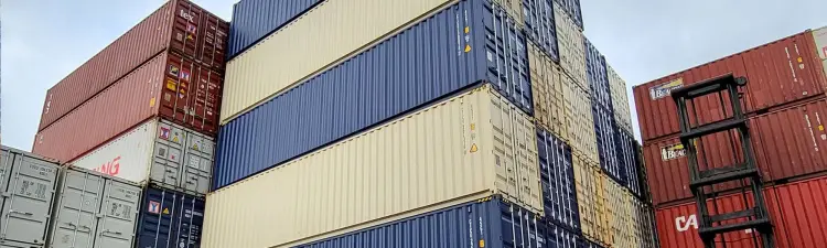 Multiple brands of shipping containers stacked on top of one another in a shipping container yard with a forklift driving by.