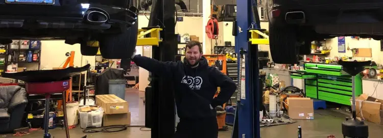 A car garage with a car lifted off the ground to be worked on. Man is leaning on the tire of the car and smiling at the camera. 