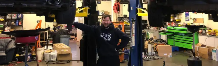 A car garage with a car lifted off the ground to be worked on. Man is leaning on the tire of the car and smiling at the camera. 