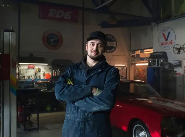 Man in blue overalls smiling with crossed arms while standing in a vehicle repair shop with a red sports car being fixed behind him.