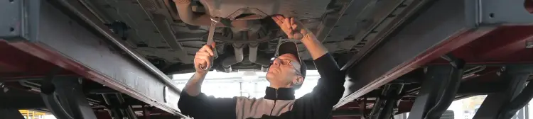 Man with baseball hat and glasses holding a wrench and working on the underside of a car while the car is up on a lift.