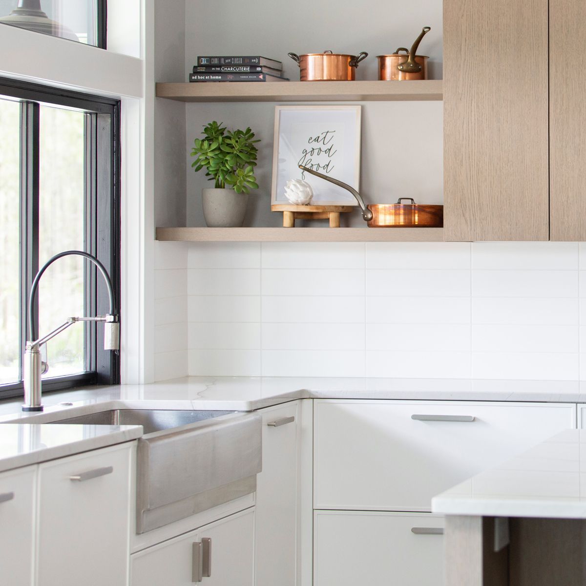 Modern kitchen with silver ranch style sink, white tile backsplash, white cabinets and countertops with accents of light-colored wood. Open-faced cabinet with copper pots