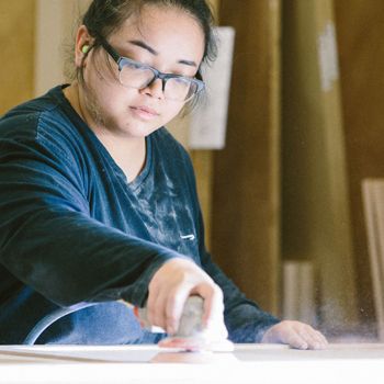 Asian woman in safety classes and a blue long-sleeved shirt sanding a wood cabinet on the table in front of her 