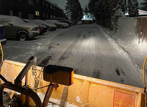 Close up of a snow plow in the early morning with its brights on as it cleans the street of a layer of snow in an apartment complex. 